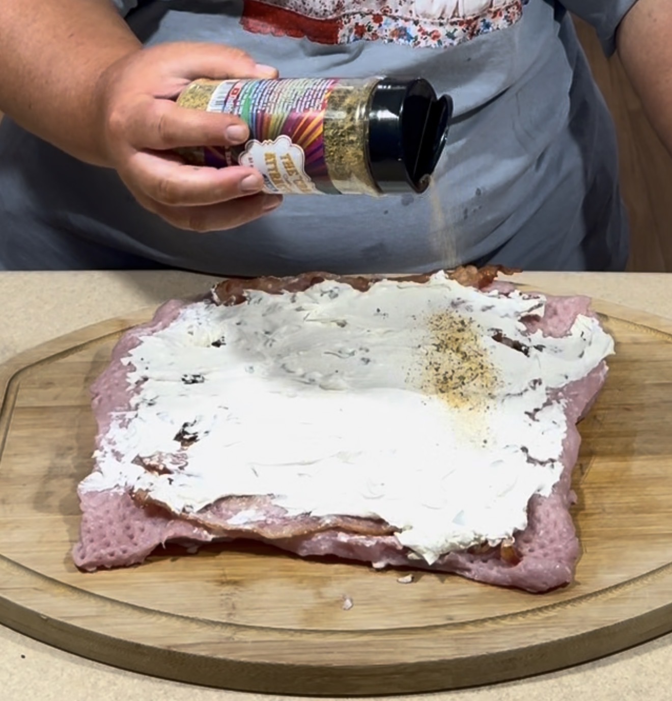 A person sprinkles seasoning onto a slab of raw meat covered with a layer of cream cheese, all placed on a wooden cutting board.