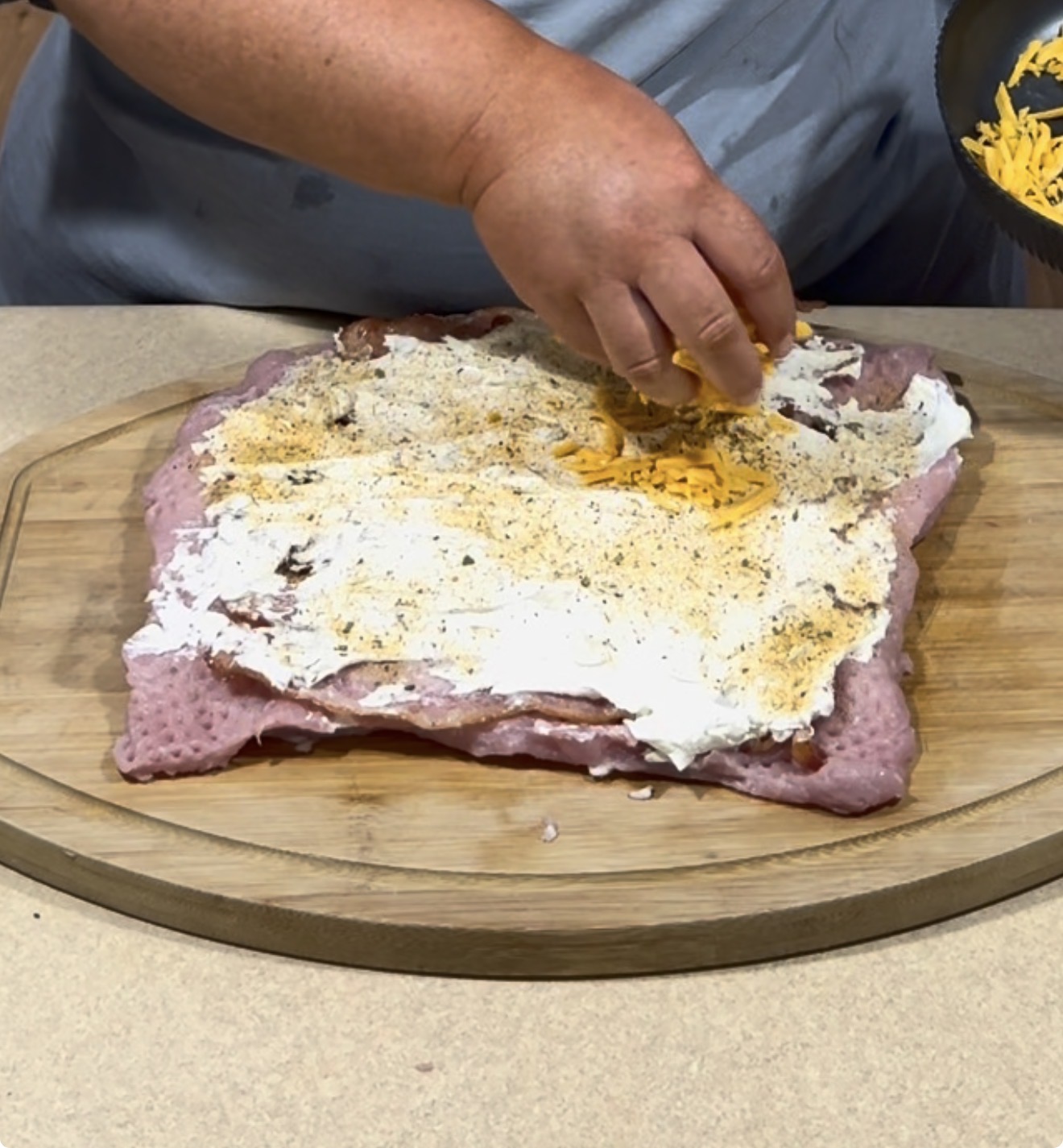A person sprinkles shredded cheese over a layer of cream cheese and seasoning spread on flattened raw turkey, which is placed on a wooden cutting board.