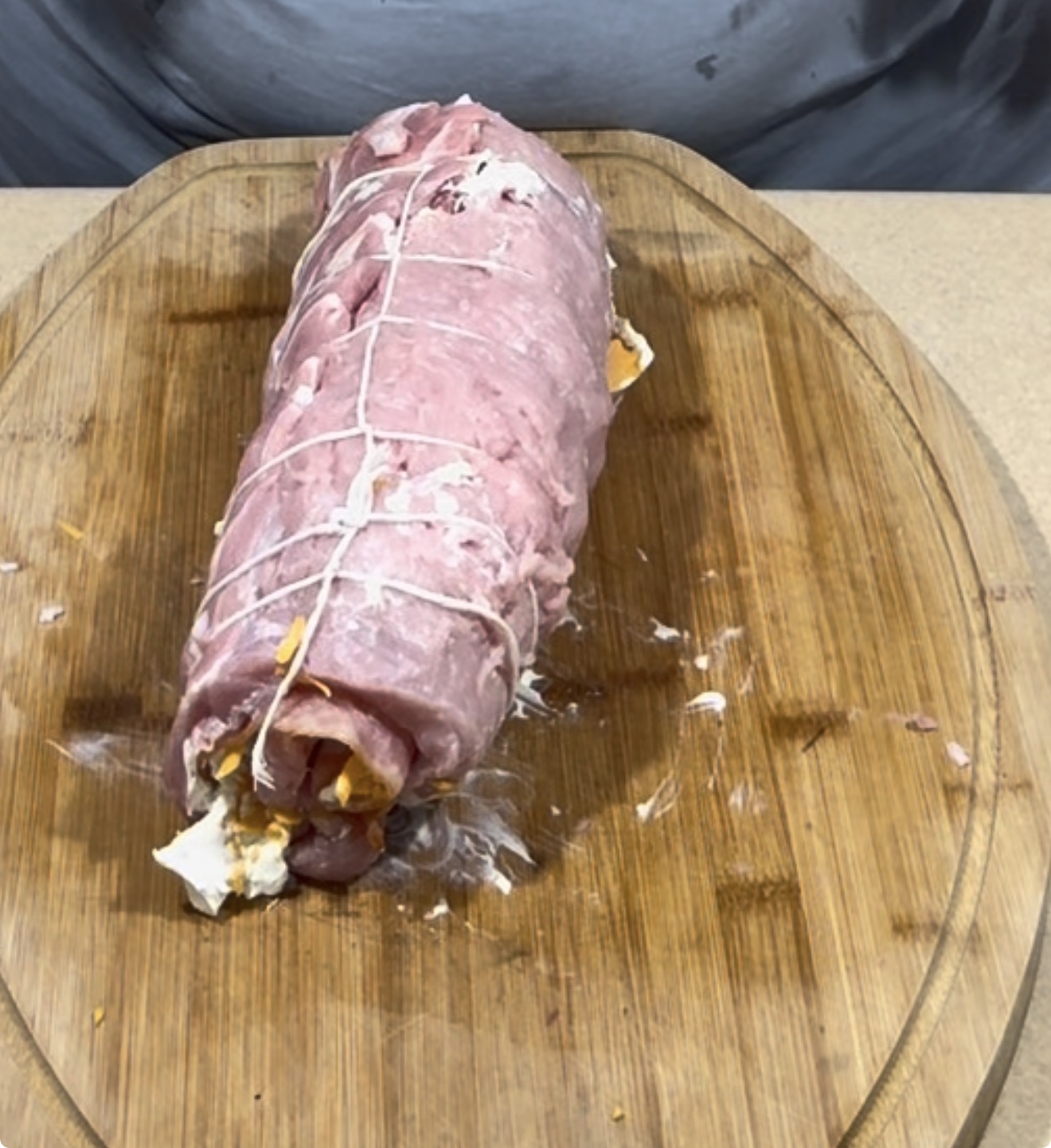 A raw, stuffed pork loin tied with kitchen twine sits on a wooden cutting board, with stuffing visible at one end. A person stands behind the board, wearing a gray shirt.