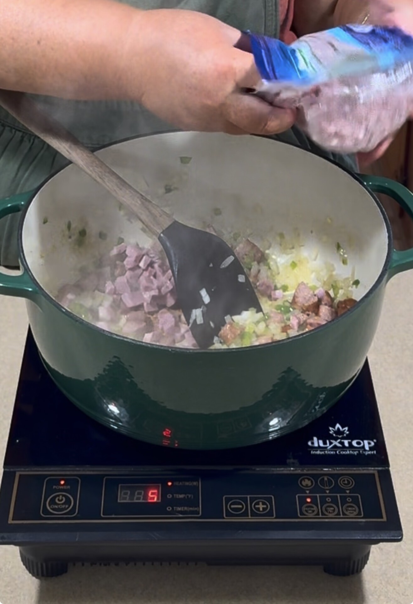 A person adds diced ham from a package into a green pot on an induction cooktop, where onions and other ingredients are being stirred with a wooden spoon. Steam rises from the cooking mixture.