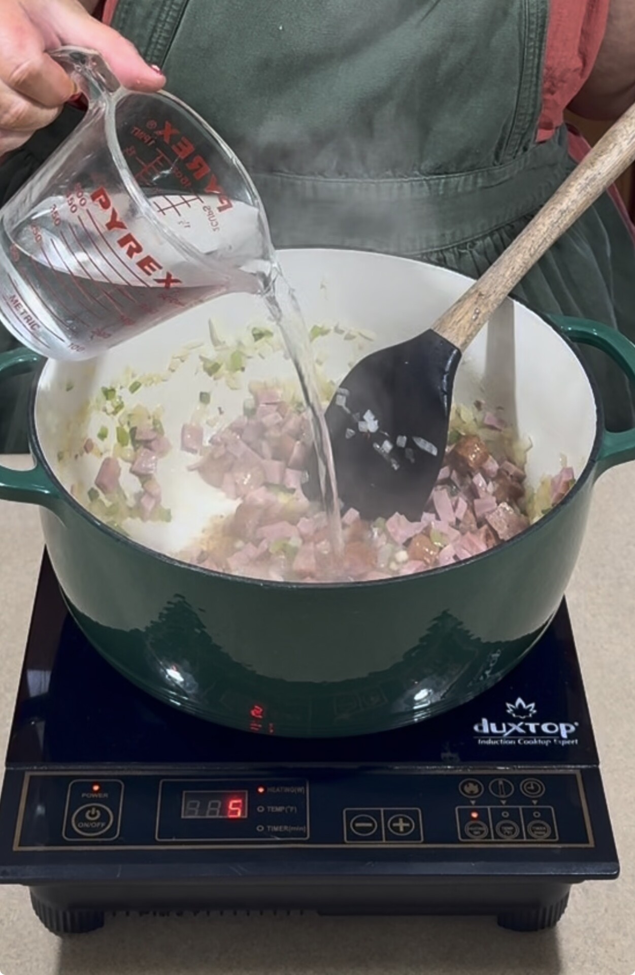 A person pours water from a measuring cup into a green Dutch oven with diced ham and onions cooking, while stirring with a black spatula on an induction cooktop.
