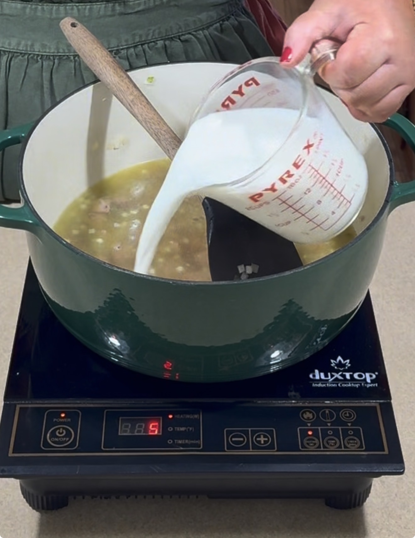 A person pours milk from a Pyrex measuring cup into a green pot of soup on an induction cooktop, stirring with a wooden spoon.
