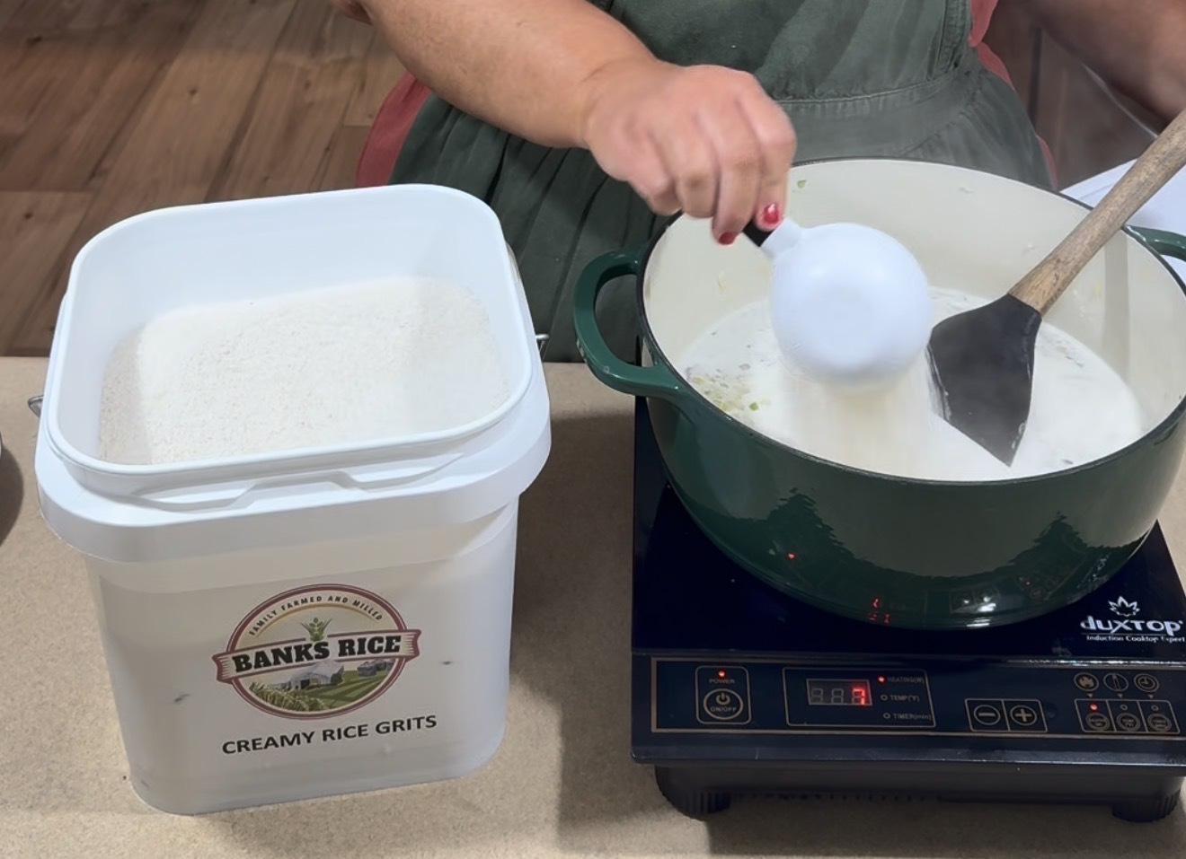 A person stirs a pot of liquid on an induction cooktop while holding a measuring cup. Next to them is a large container labeled Banks Rice Creamy Rice Grits, containing white grits.