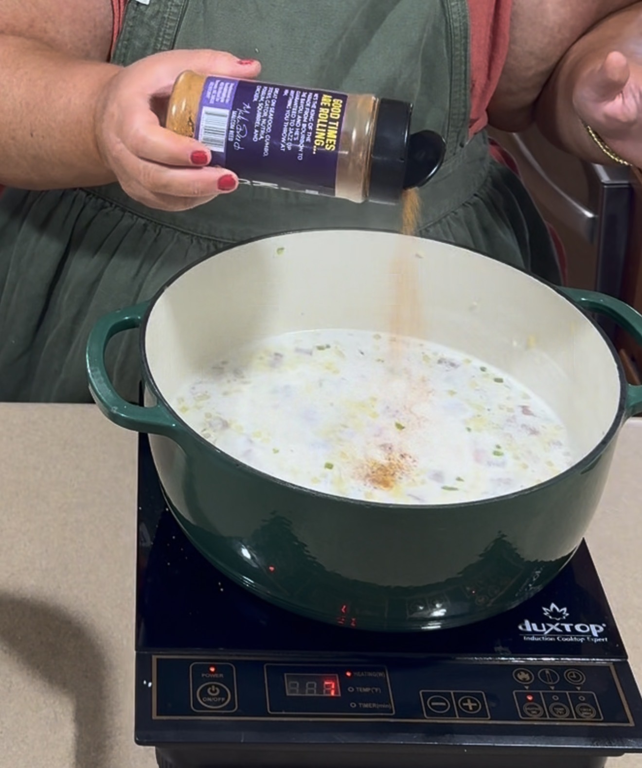 A person sprinkles seasoning from a jar into a green Dutch oven filled with a creamy mixture, cooking on an induction stovetop.