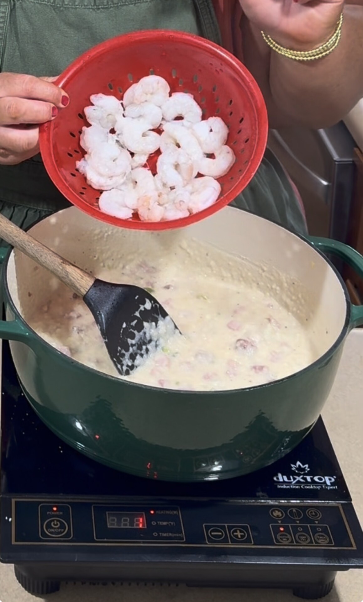 A person holding a red colander of raw shrimp over a green pot of creamy soup on an induction stove, preparing to add the shrimp to the dish. A black spatula rests inside the pot.