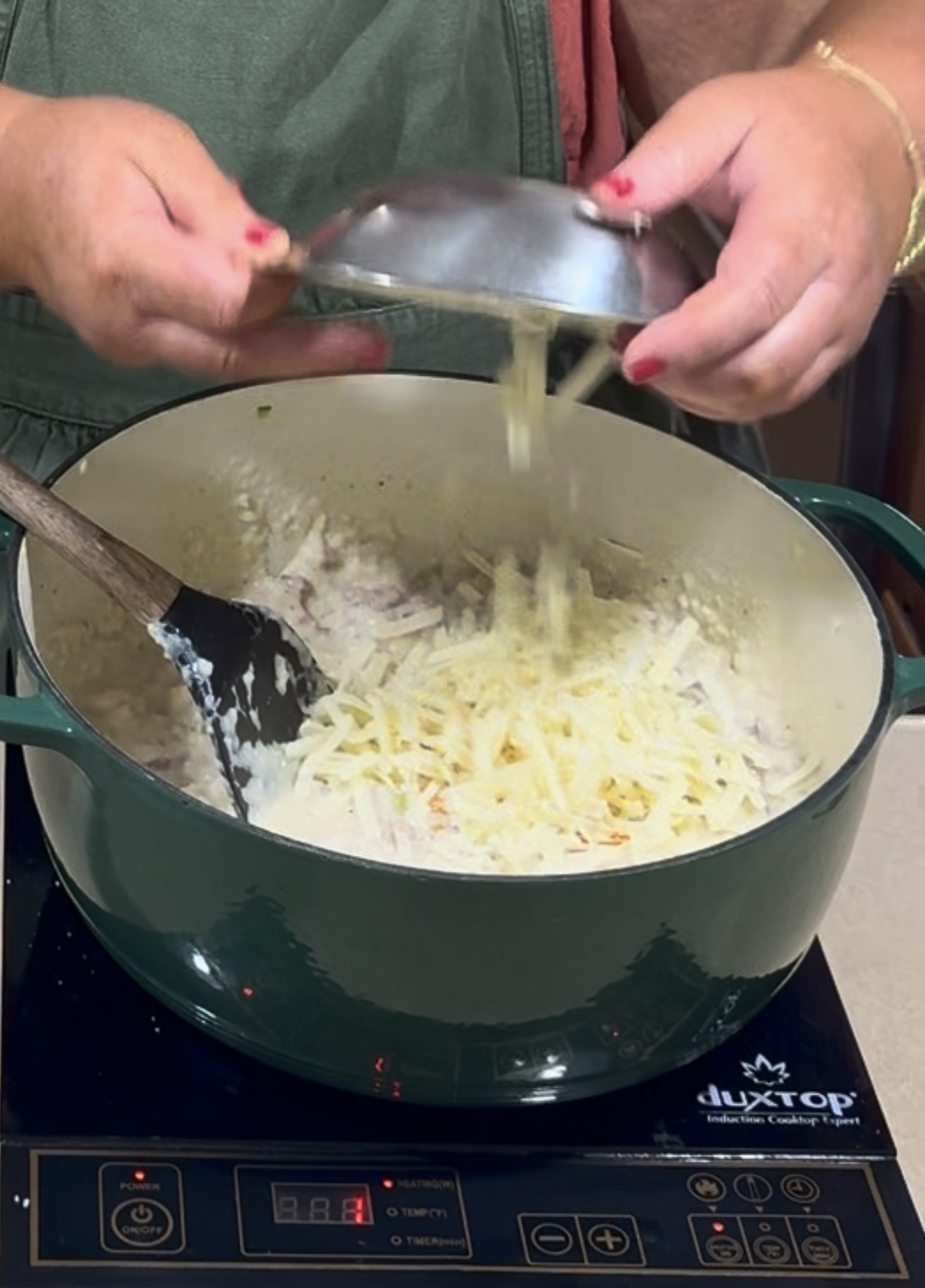 A person adds shredded cheese from a metal bowl into a green Dutch oven on an electric stovetop, stirring a creamy mixture with a black spatula.