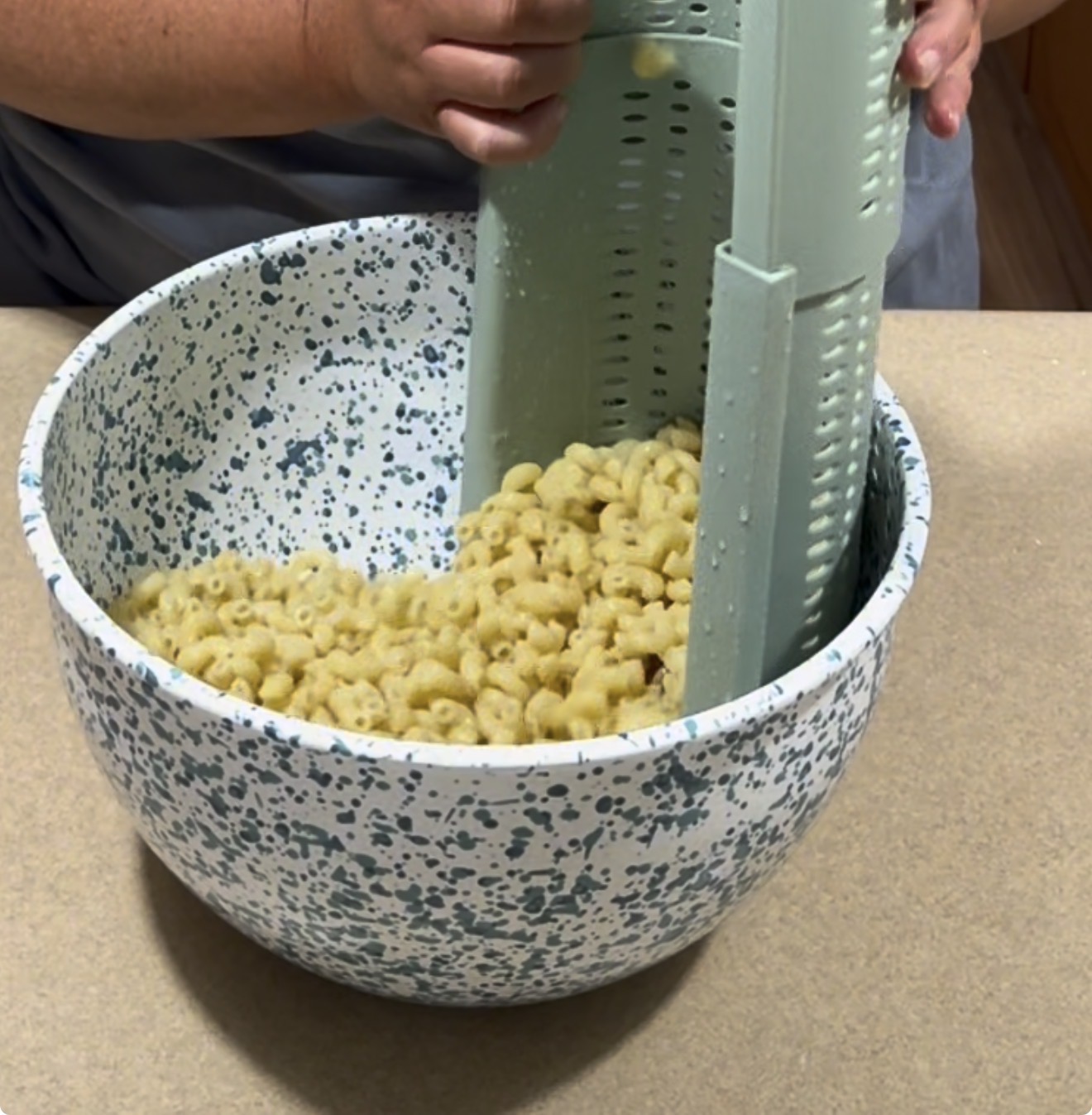 A person drains cooked macaroni pasta using a vertical strainer inside a large speckled mixing bowl on a countertop.