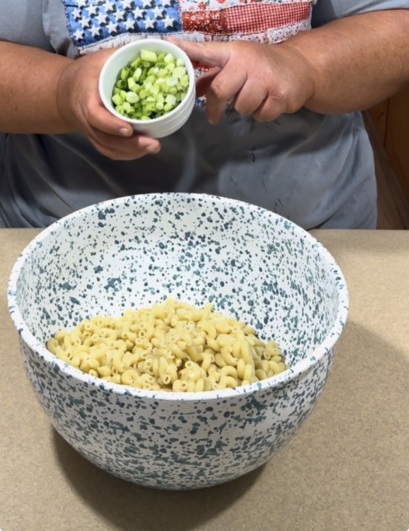 A person holding a small bowl of chopped green onions above a large speckled mixing bowl filled with cooked elbow macaroni on a countertop.