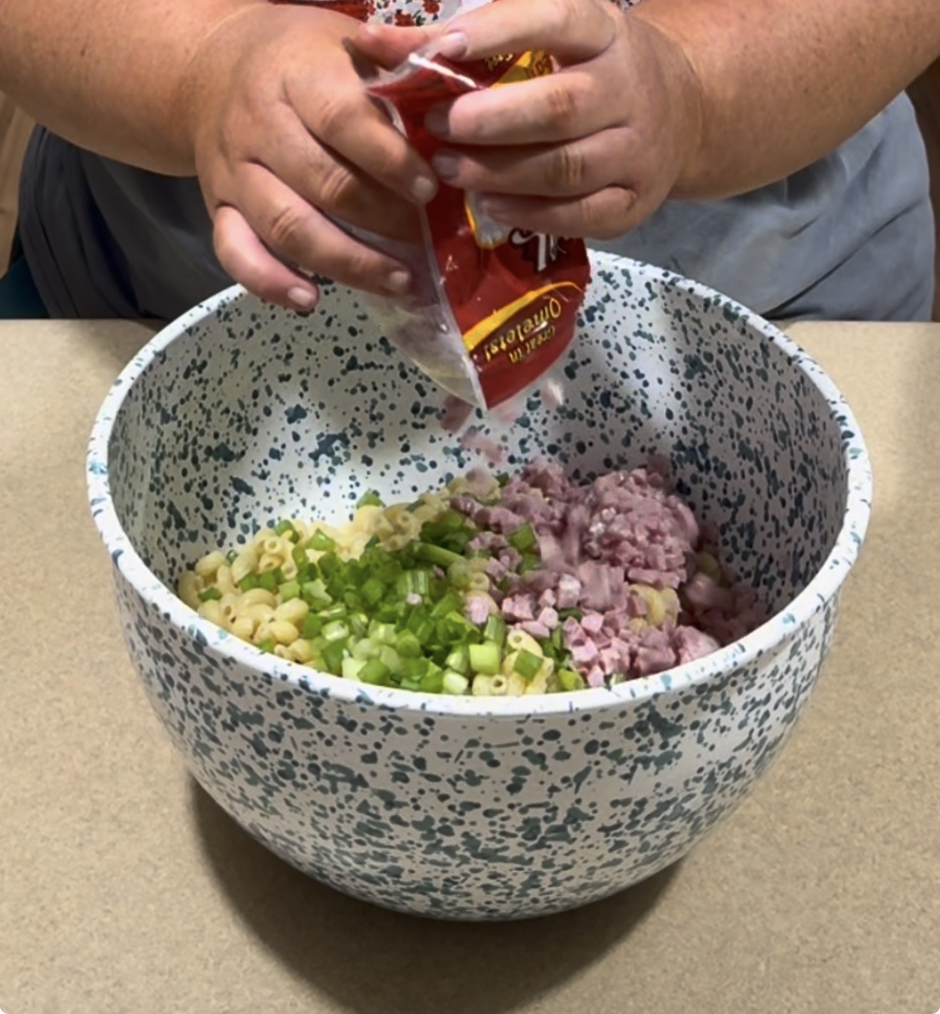 A person pours shredded cheese from a bag into a speckled mixing bowl containing chopped celery, diced ham, and elbow macaroni on a kitchen counter.