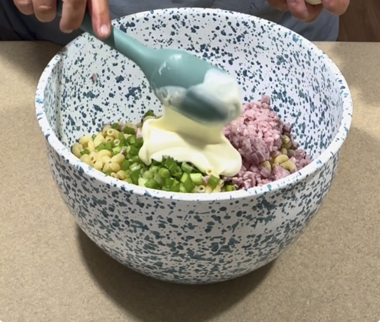 A person holds a blue spatula with mayonnaise over a speckled mixing bowl filled with cooked pasta, chopped ham, green bell pepper, and peas on a countertop.