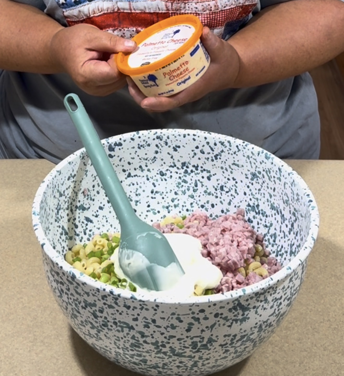 A person holds a container of Palmetto Cheese above a speckled mixing bowl filled with chopped ham, pasta, green onions, and a dollop of creamy dressing. A teal spatula rests inside the bowl.