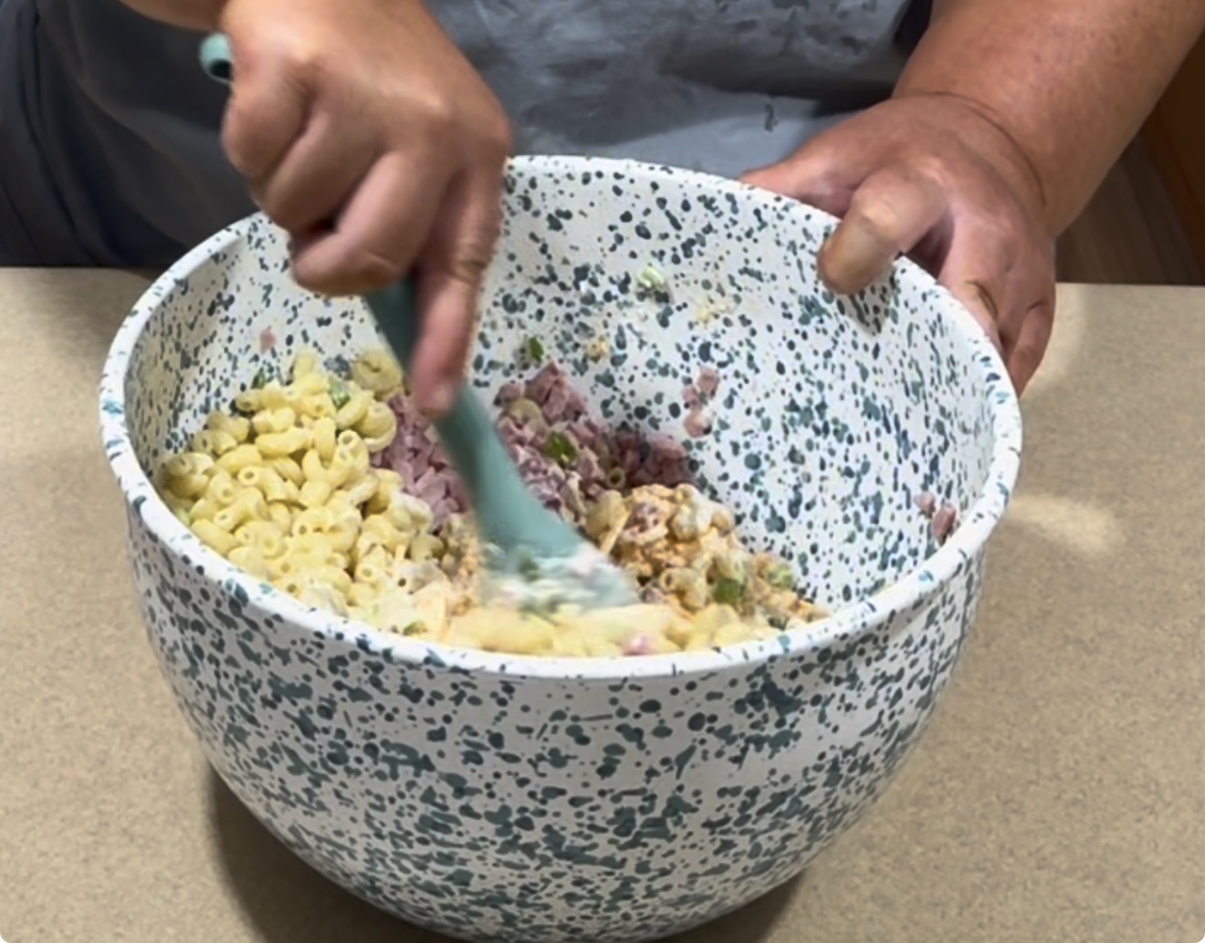 A person is mixing elbow macaroni, diced ham, and other ingredients in a large, speckled bowl with a green spatula on a kitchen countertop.