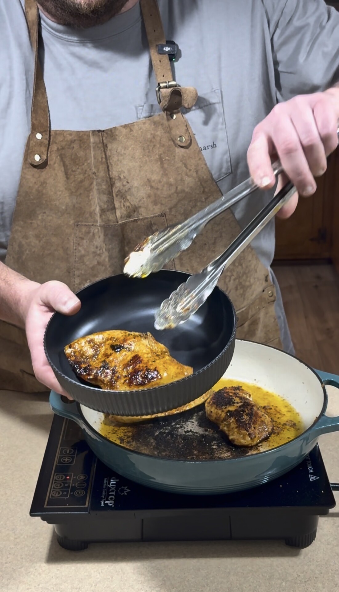 A person wearing a brown apron uses metal tongs to place a cooked, seasoned chicken breast from a pan on the stove into a black bowl.