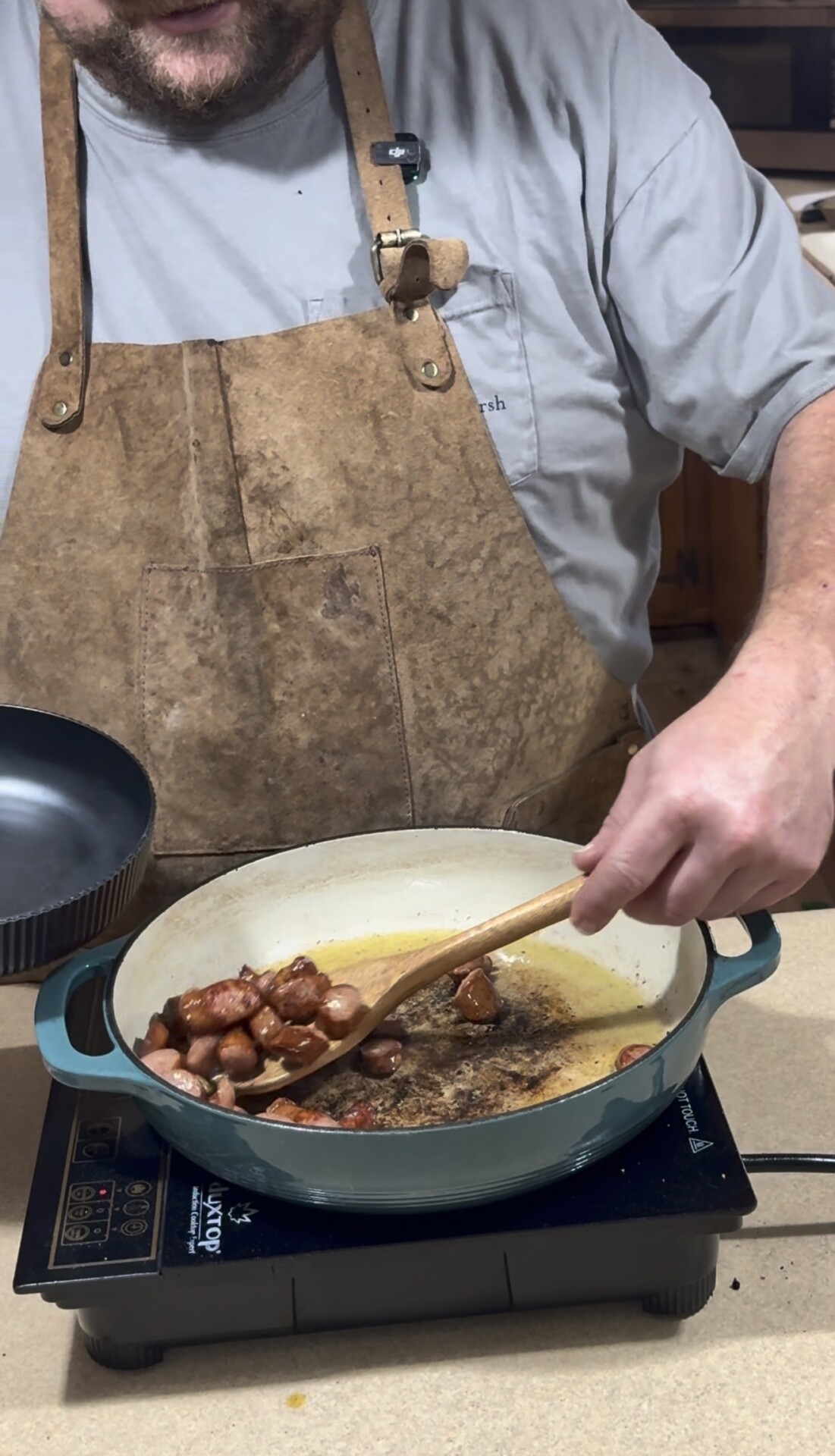 A person wearing a brown apron is cooking sliced sausage pieces in a large pot on a stove, stirring them with a wooden spoon while holding a dark bowl in the other hand.