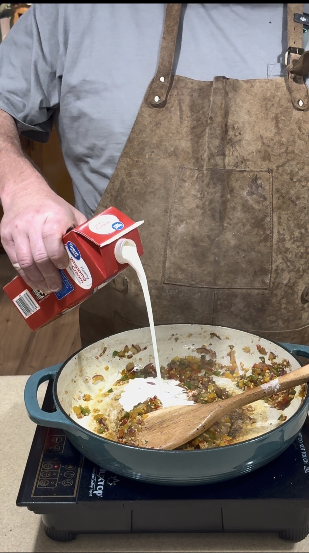 A person wearing a brown apron pours heavy cream from a red carton into a pan of cooked vegetables on a stovetop, stirring with a wooden spoon.