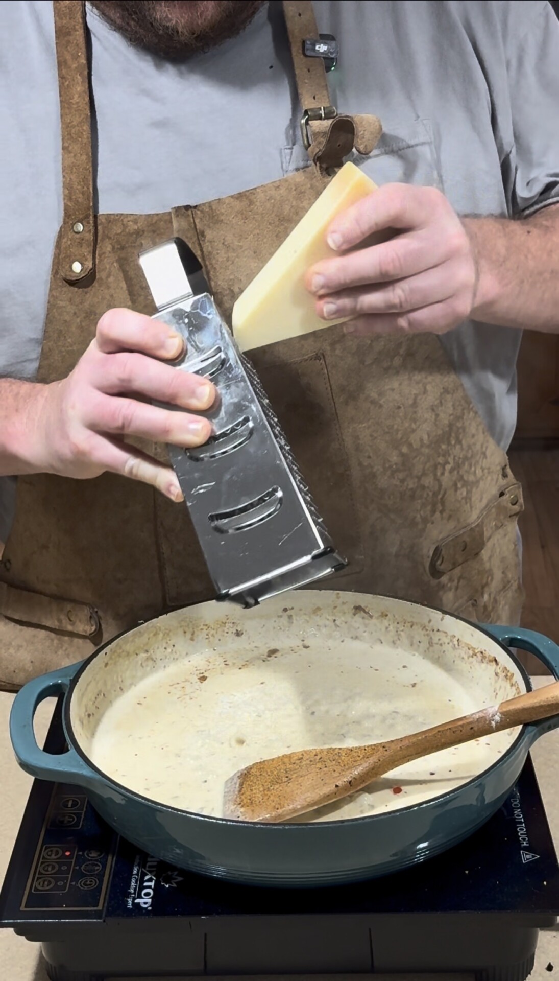 A person in a brown apron grates a block of cheese over a creamy sauce in a blue pot on a stove, with a wooden spoon resting inside the pot.