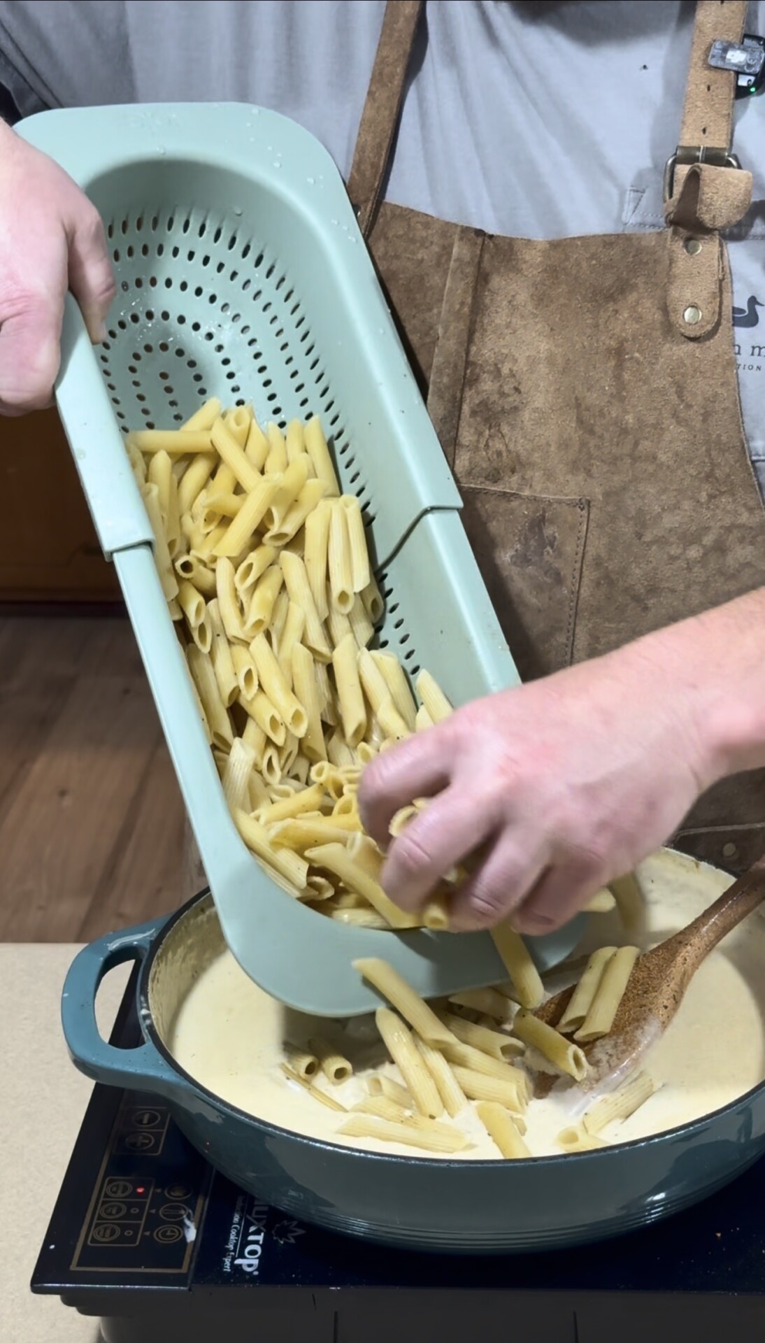 A person wearing a brown apron pours cooked penne pasta from a teal colander into a pot of creamy sauce on a stovetop, using a wooden spoon to stir.