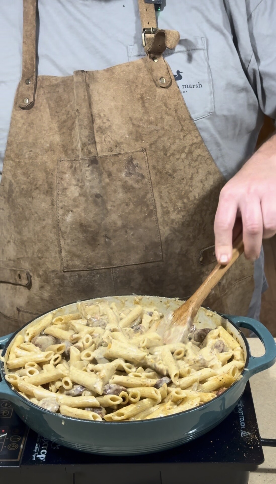 A person wearing a brown apron is stirring a large pan filled with creamy pasta and pieces of meat or mushrooms using a wooden spoon. The pan is on a stovetop.