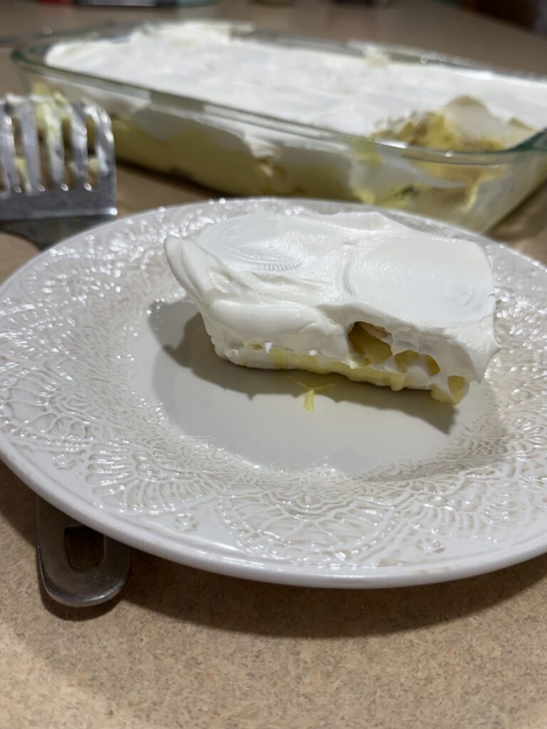 A slice of a creamy dessert with a white topping sits on a decorative white plate. The rest of the dessert is in a glass baking dish in the background, with a serving spatula nearby.