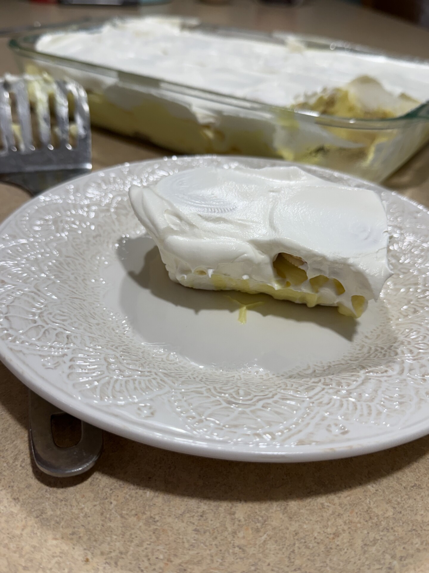 A slice of a creamy dessert with a white topping sits on a decorative white plate. The rest of the dessert is in a glass baking dish in the background, with a serving spatula nearby.