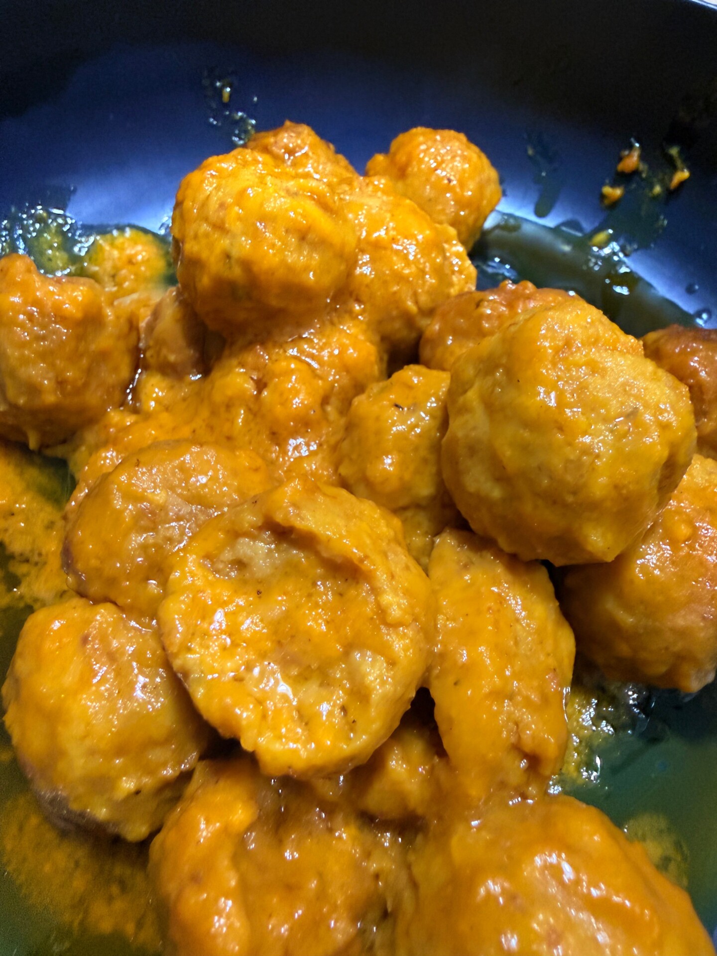 A close-up of several golden-brown meatballs coated in a thick orange sauce, arranged in a dark-colored bowl.