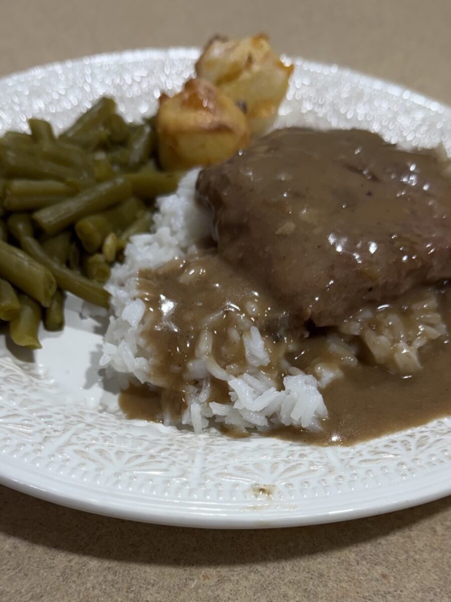 A white plate holds green beans, two small roasted potatoes, and white rice topped with a brown gravy-covered meat patty, all placed on a beige surface.