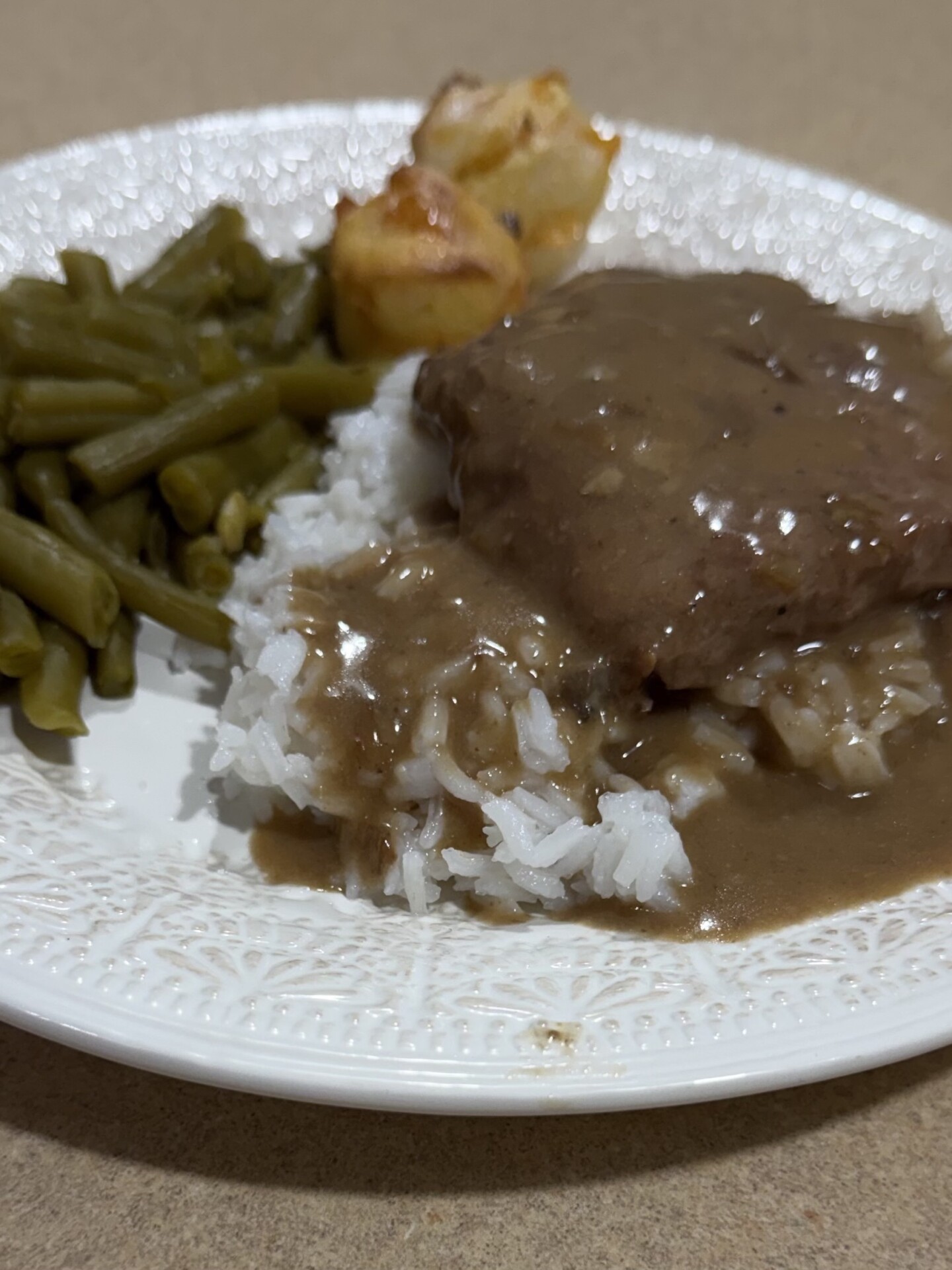 A white plate holds green beans, two small roasted potatoes, and white rice topped with a brown gravy-covered meat patty, all placed on a beige surface.