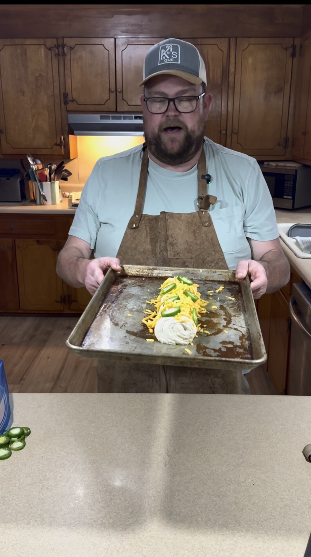 A man wearing glasses, a cap, and an apron stands in a kitchen holding a baking tray with a rolled dough topped with shredded cheese and green peppers. Wooden cabinets, a stove, and a counter are visible in the background.