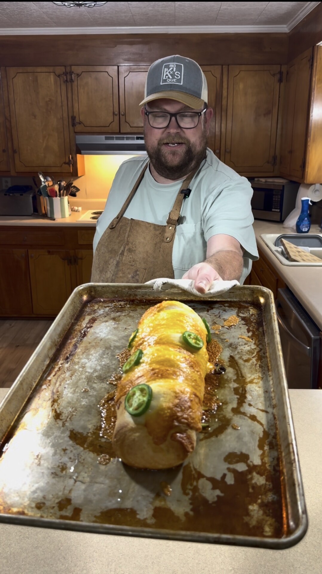 A man in a kitchen, wearing glasses, a cap, and an apron, holds out a baking tray with a golden, cheesy loaf topped with sliced jalapeños. Wooden cabinets and kitchen utensils are in the background.