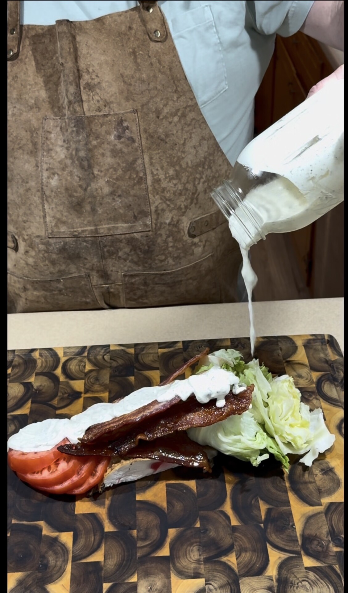 A person wearing brown overalls pours dressing from a jar onto a wedge salad with bacon, tomato, and lettuce on a wooden cutting board.