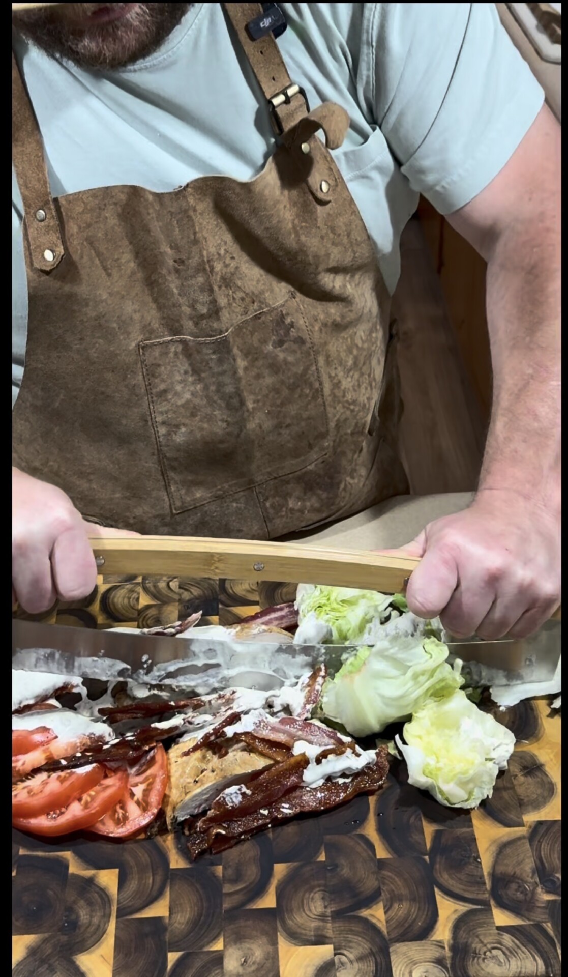 A person wearing a brown apron slices cooked meat on a wooden cutting board with a large knife. Sliced tomatoes, lettuce, and a drizzle of white sauce are arranged beside the meat.