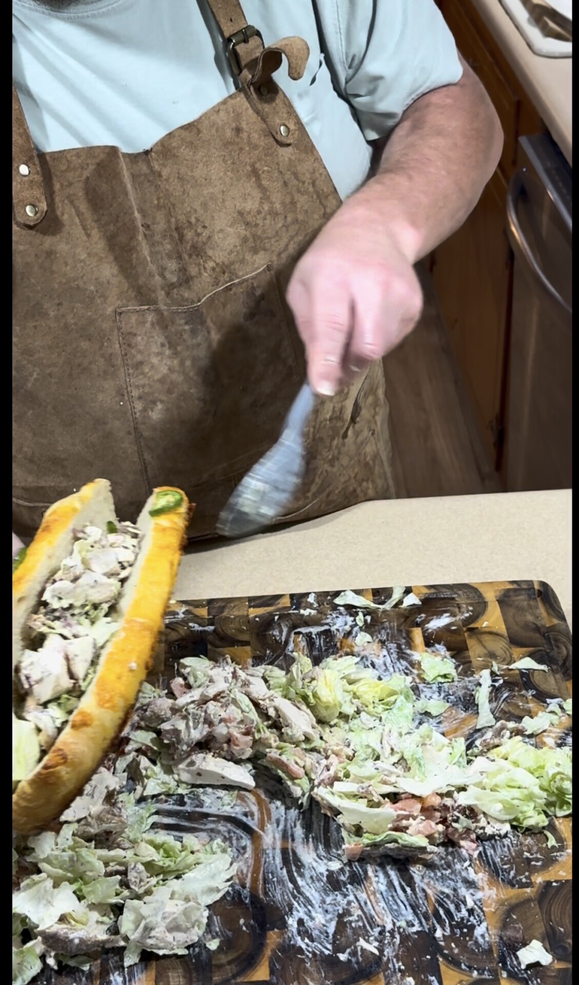 A person wearing a brown apron is making a sandwich on a wooden cutting board, filling a baguette with shredded meat, lettuce, and mayonnaise.