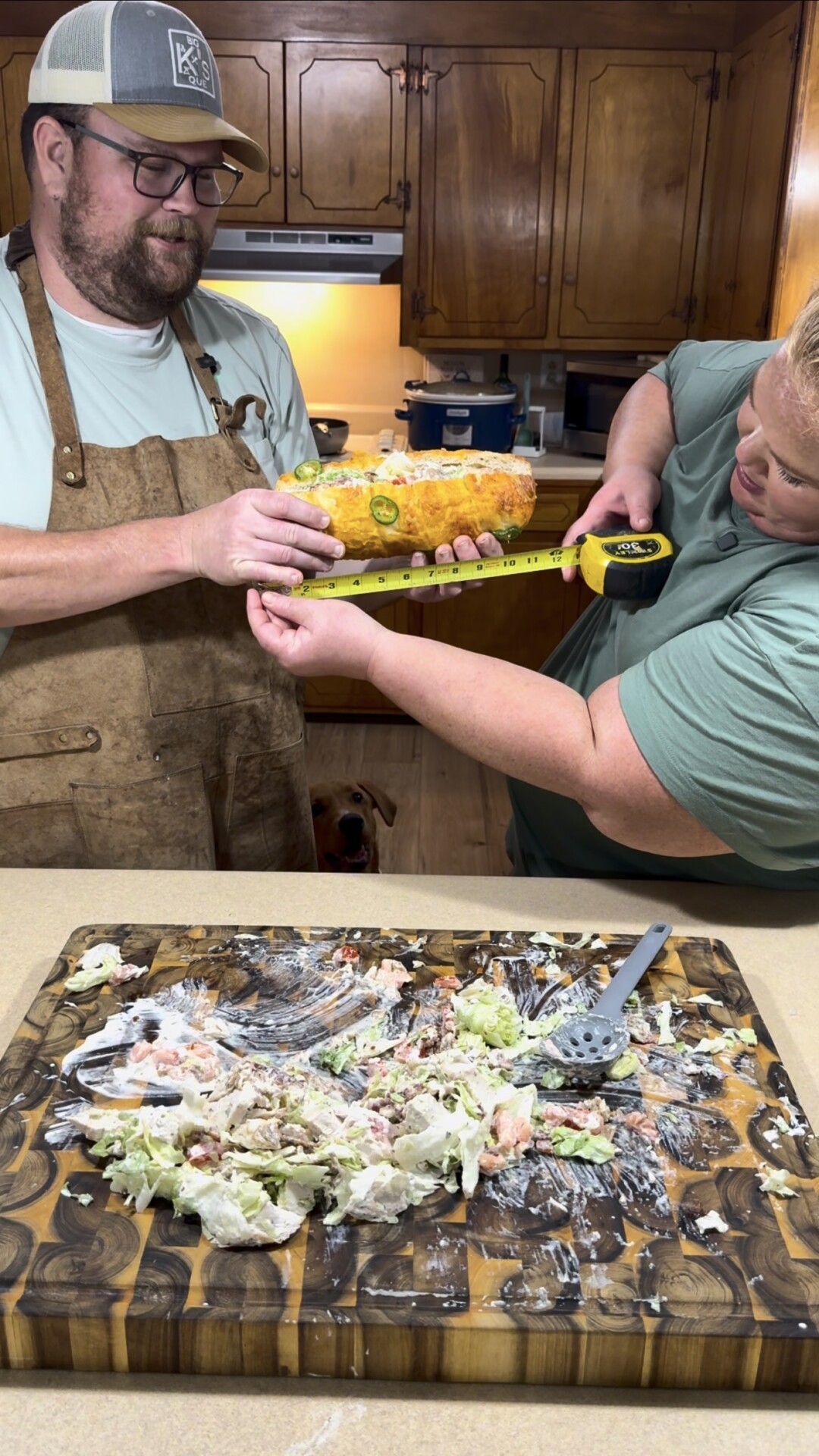 A man wearing an apron holds a large sub sandwich while a woman measures it with a tape measure. In front of them, a cutting board holds the remains of a mixed salad. The scene is set in a kitchen.