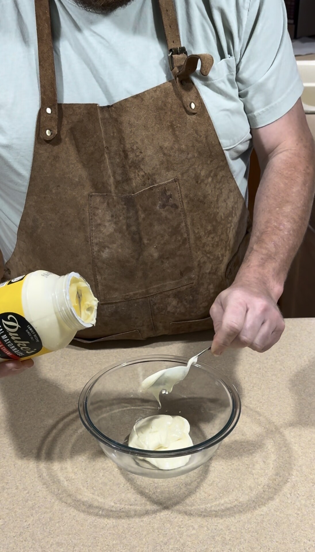 A person wearing a brown apron transfers mayonnaise from a jar into a clear glass bowl using a spoon, standing at a kitchen counter.