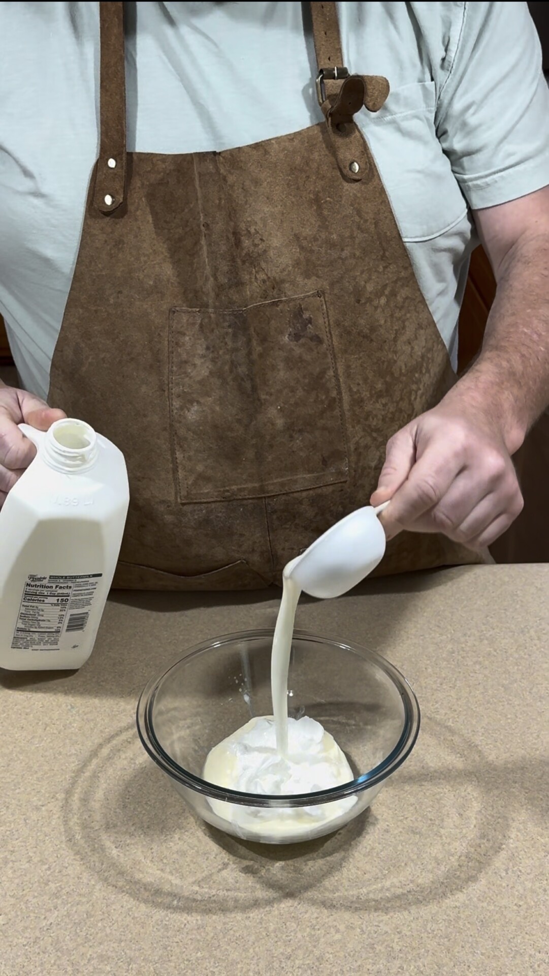 A person wearing a brown apron is pouring milk from a jug into a glass bowl containing a white creamy substance on a kitchen counter.