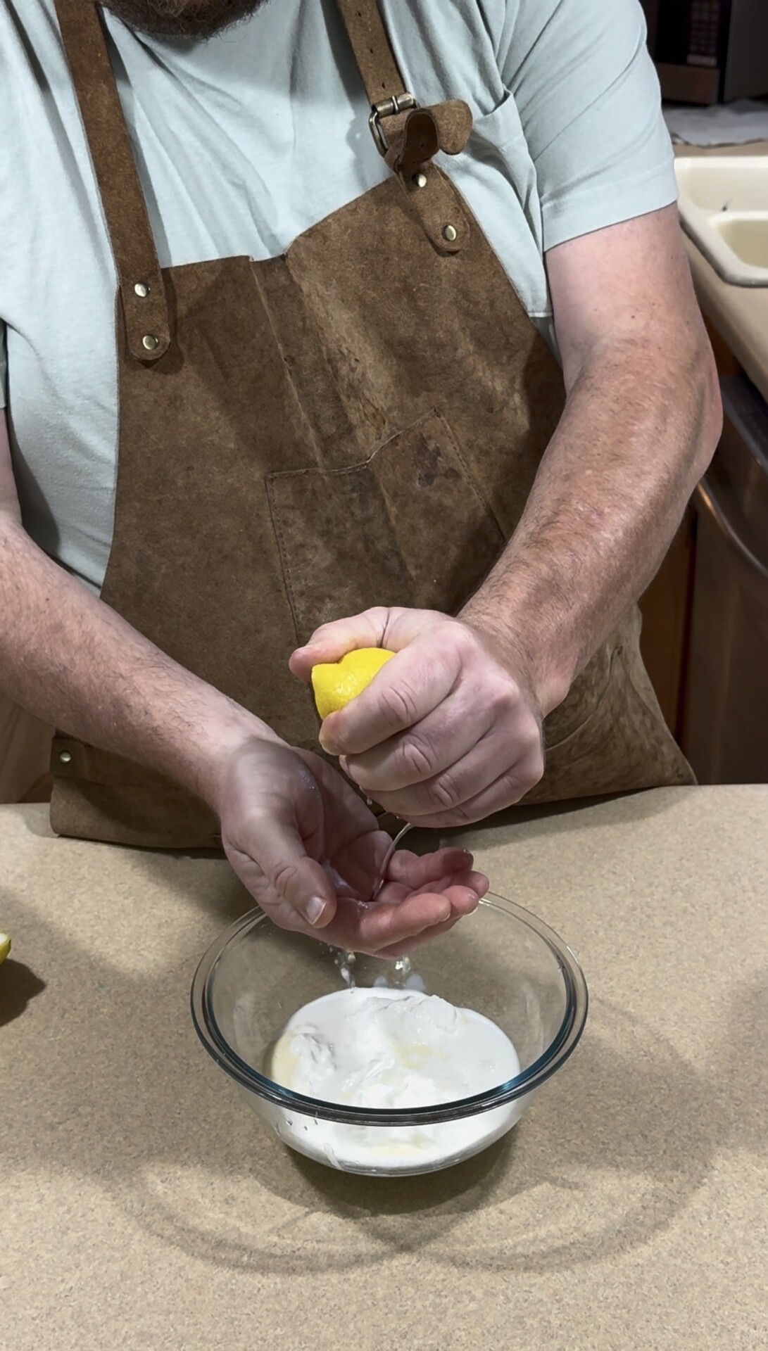 A person wearing a brown apron squeezes half a lemon over a glass bowl filled with white yogurt or sour cream on a kitchen counter.