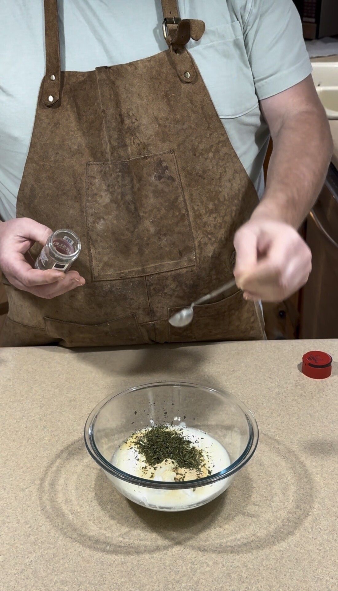 A person wearing a brown apron is adding dried herbs from a small jar into a glass bowl containing a creamy mixture on a kitchen counter.