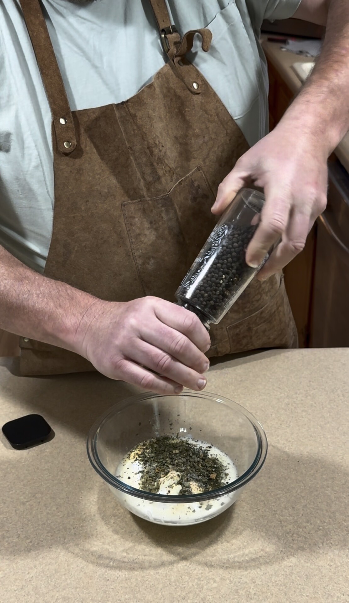 A person wearing a brown apron grinds black pepper into a glass bowl filled with herbs and a creamy mixture, standing at a kitchen counter.