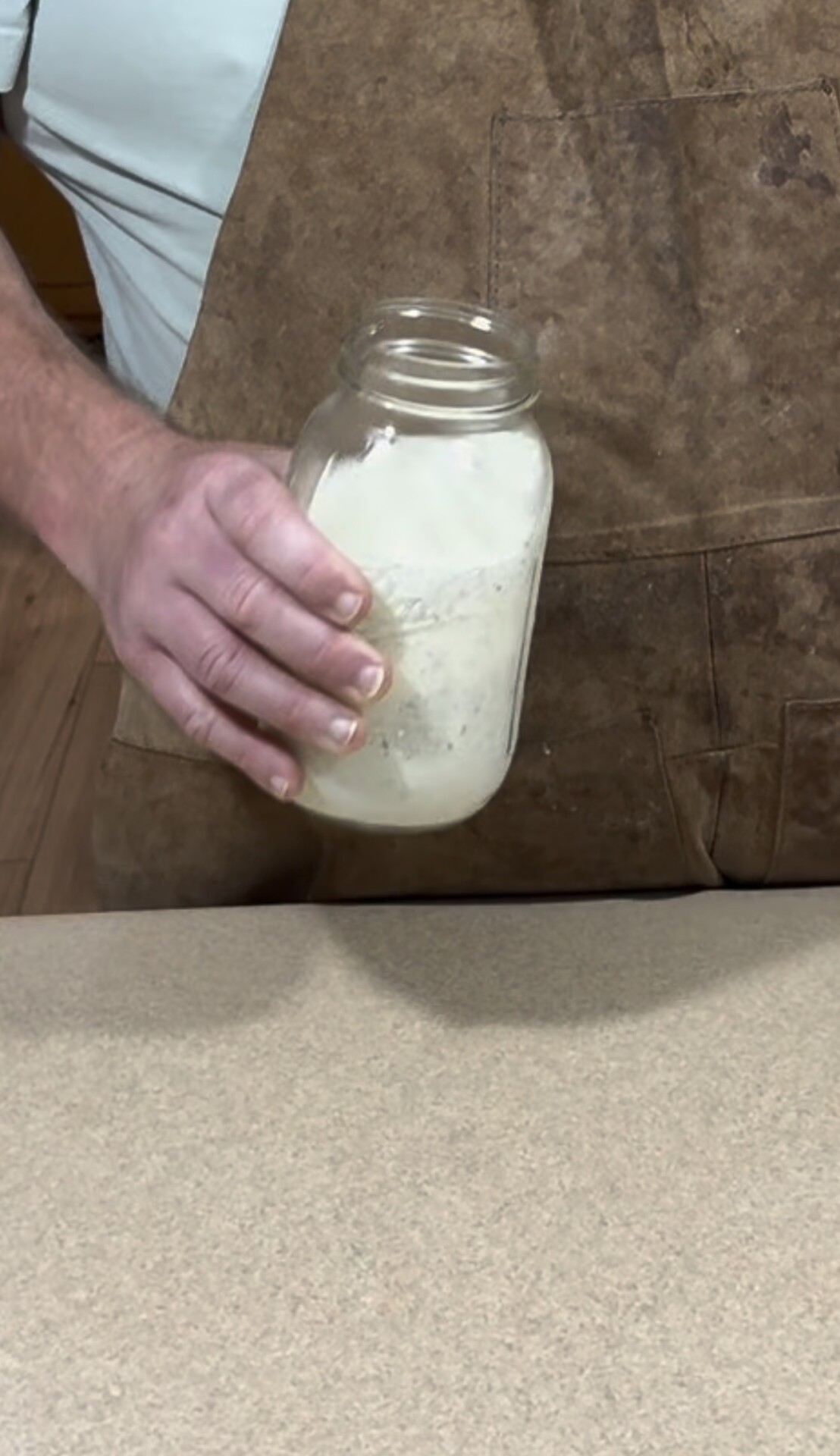 A person wearing a brown apron holds a glass jar containing a white powdery substance, positioned above a light-colored countertop.