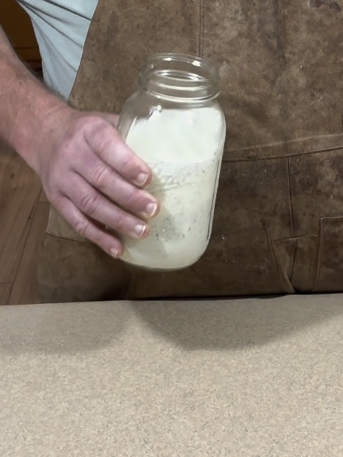 A person in a brown apron holds a glass jar containing a white, creamy substance over a countertop.