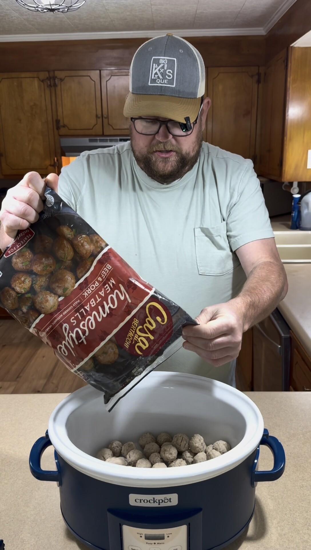 A man wearing a cap and glasses pours frozen meatballs from a bag into a blue crockpot on a kitchen counter, with wooden cabinets and a sink visible in the background.