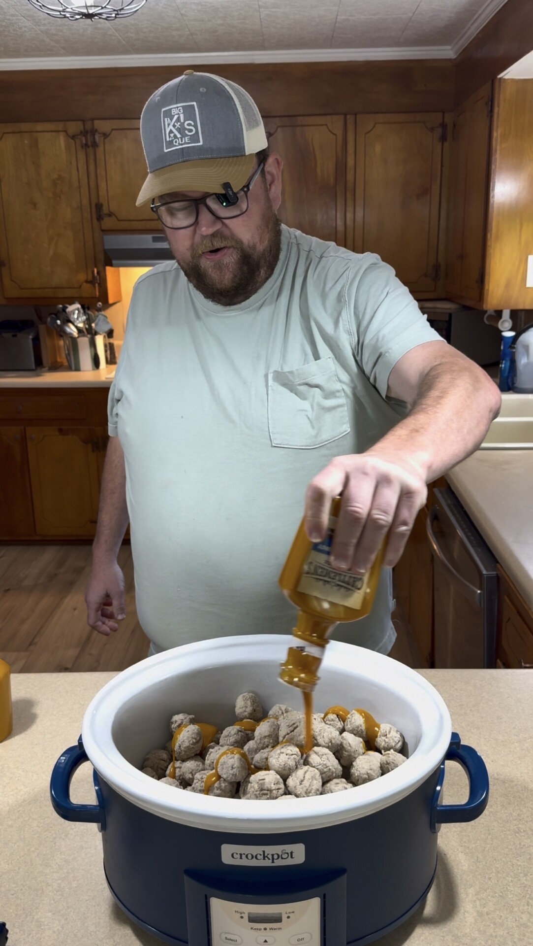 A man wearing glasses and a cap is pouring sauce from a bottle onto frozen meatballs in a slow cooker in a kitchen with wood cabinets.