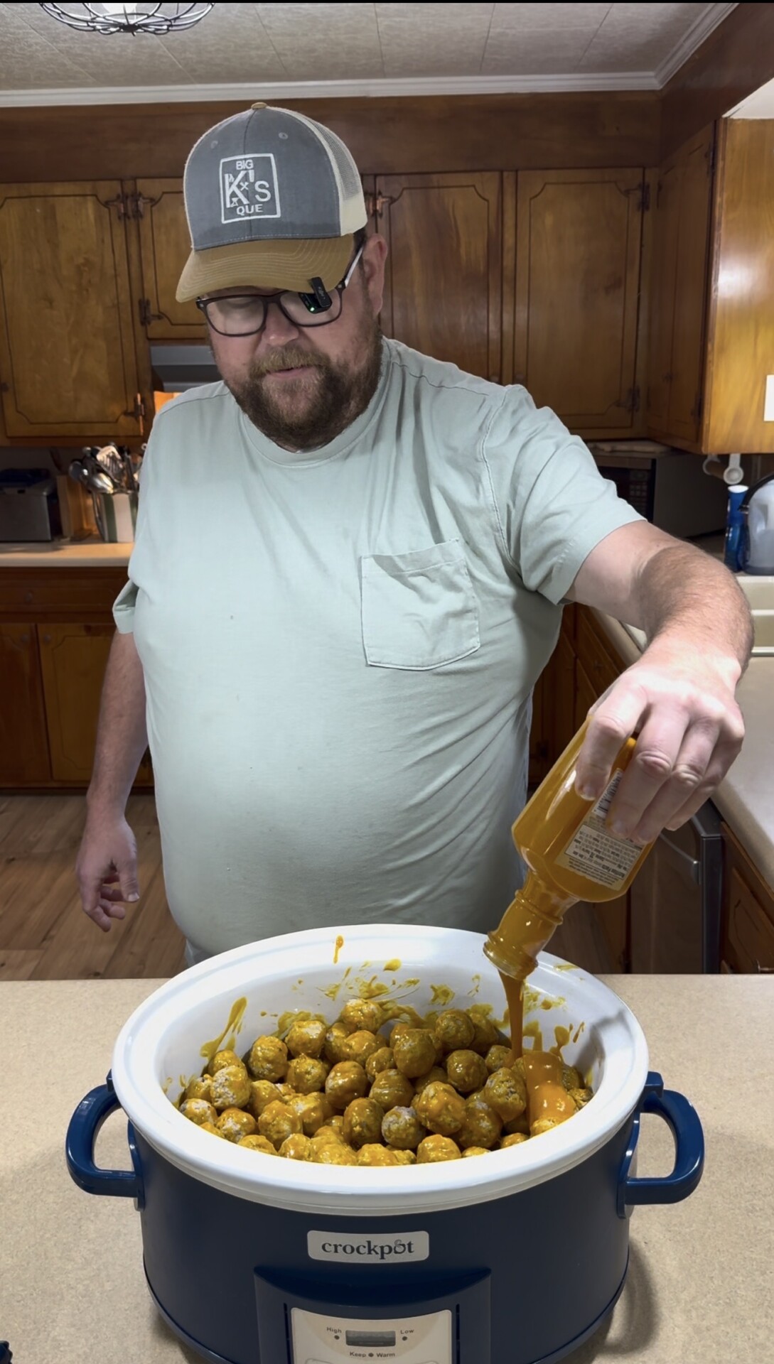 A man in a light blue shirt and cap pours yellow sauce from a bottle onto meatballs in a white Crockpot slow cooker, standing in a wood-paneled kitchen.