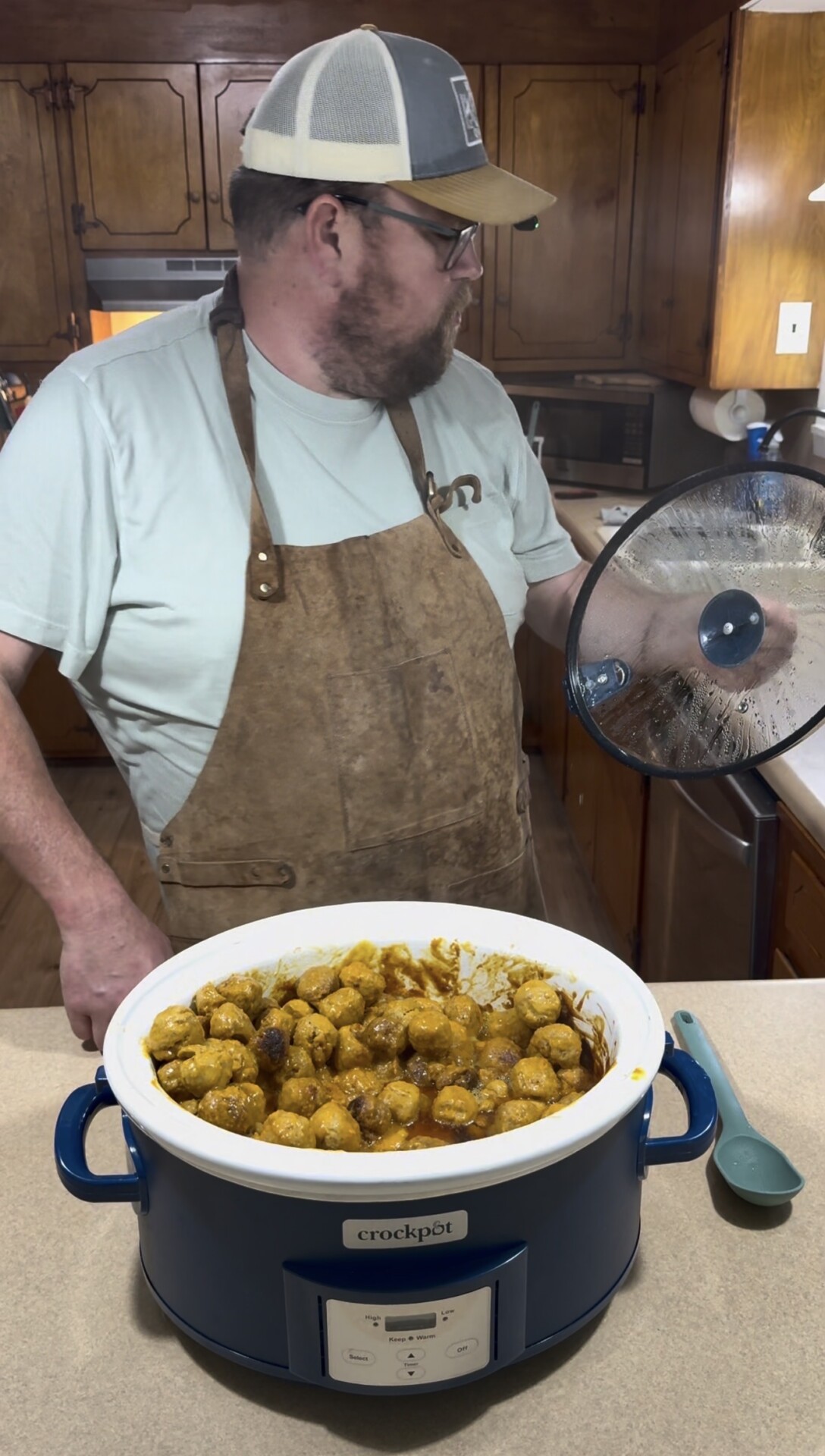 A man wearing a cap, glasses, t-shirt, and apron stands in a kitchen holding a slow cooker lid. In front of him, a crockpot filled with meatballs in sauce sits on the counter next to a blue spoon.