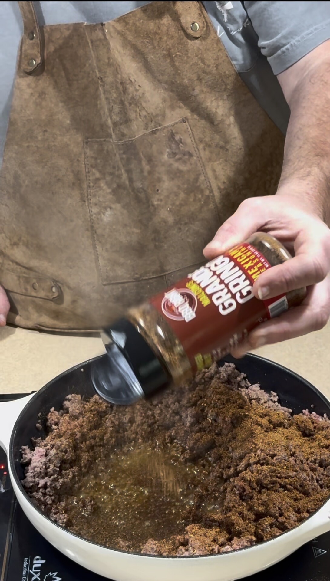 A person wearing a brown apron is seasoning cooked ground meat in a pan with a large container of Grand Grub BBQ seasoning.