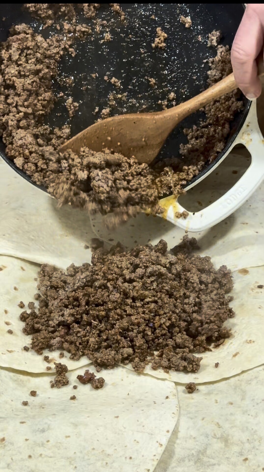 Cooked ground beef is being poured from a pan onto a flour tortilla, with a wooden spoon aiding in transferring the meat. The tortilla is laid flat on a light-colored surface.