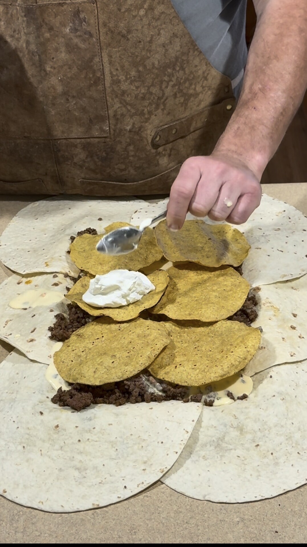 A person spreads sour cream onto a layer of hard taco shells, ground beef, and cheese, which are placed on top of overlapping flour tortillas on a countertop.