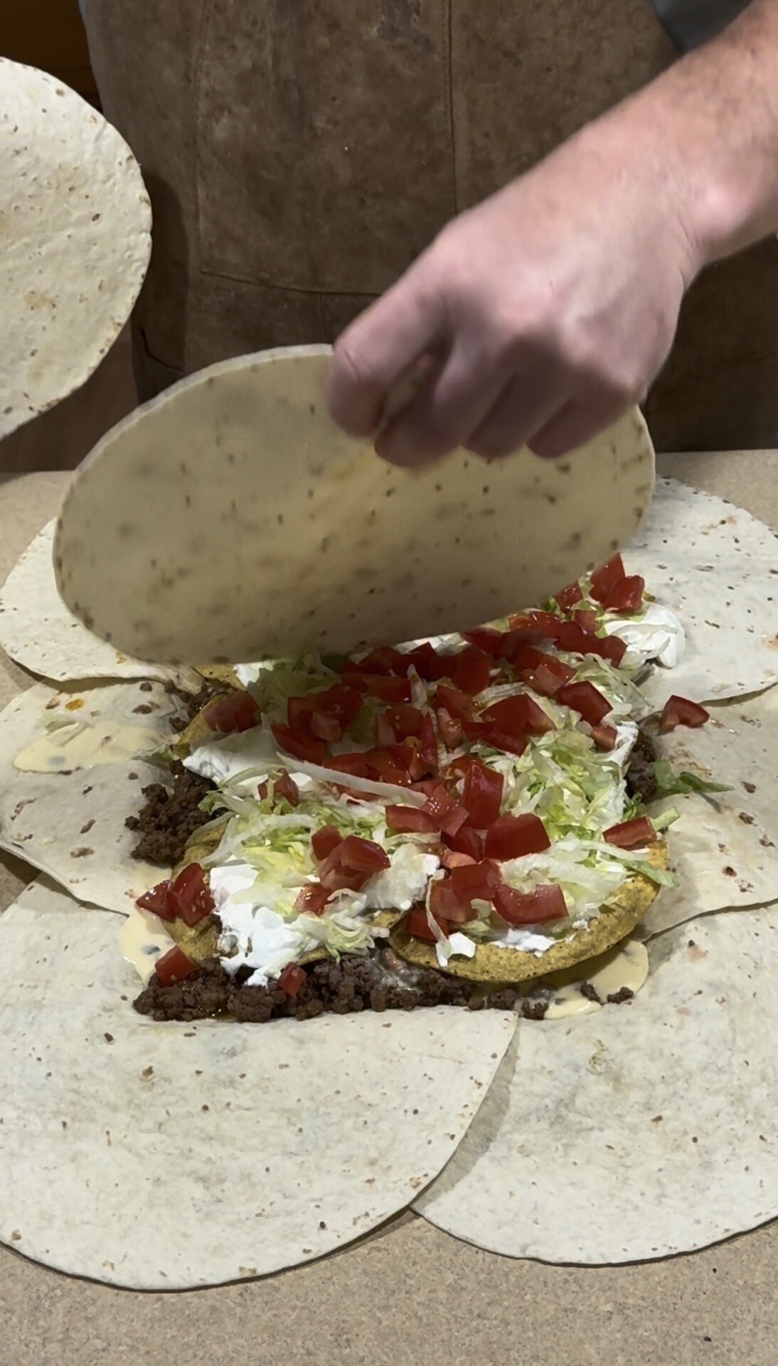A hand is folding a tortilla over a large tortilla wrap filled with ground meat, shredded lettuce, diced tomatoes, sour cream, and cheese on a flat surface, surrounded by overlapping tortillas.