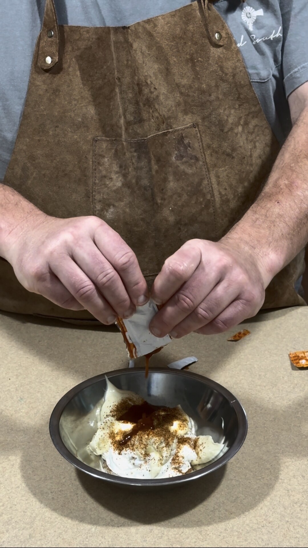 A person wearing a brown apron squeezes sauce from a small packet into a metal bowl containing a white creamy mixture and spices on a beige countertop.