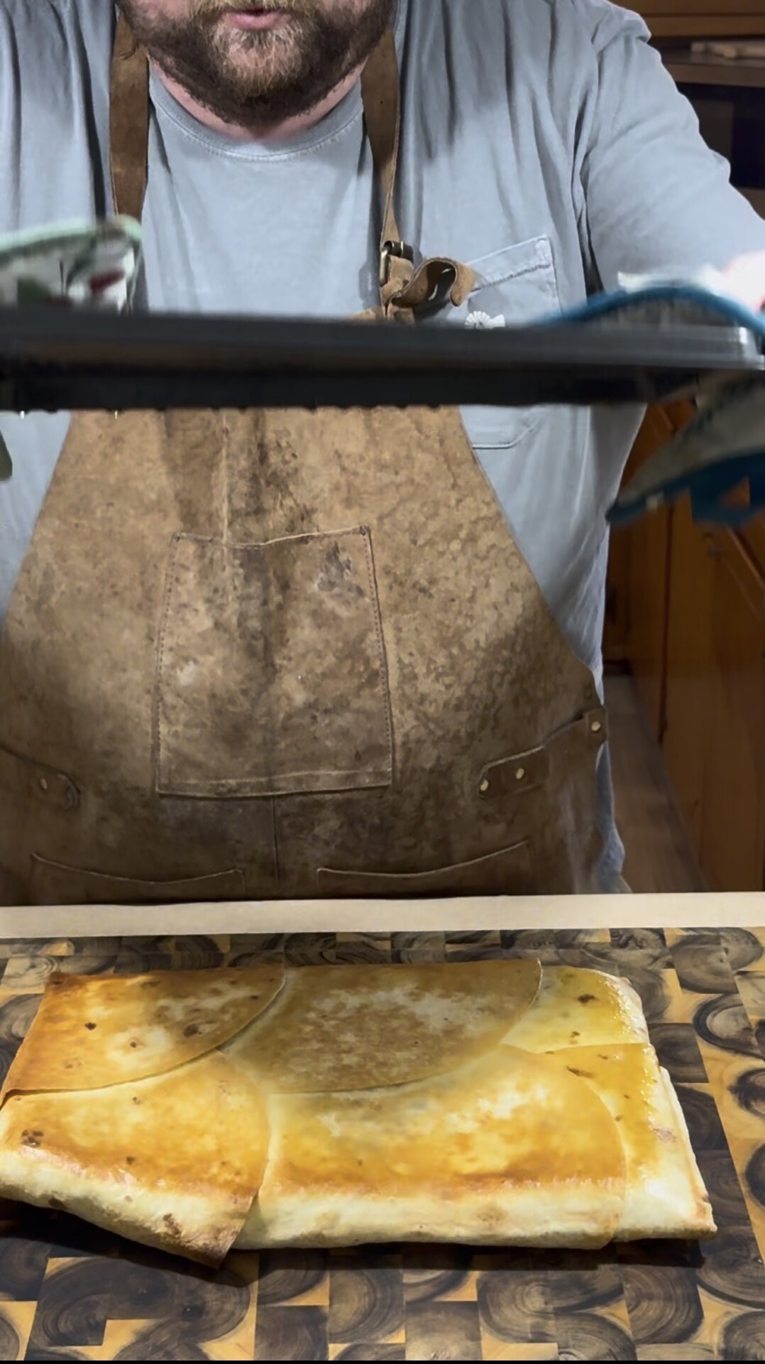 A person wearing a brown apron and gray shirt sets down a large rectangular, golden-brown baked pastry or pie onto a wooden cutting board. The persons face is not visible.